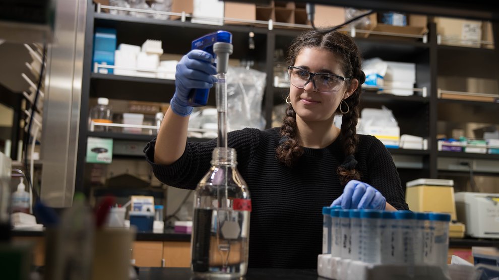 Gabby, in a lab, wearing goggles and gloves to extract liquid from a bottle.