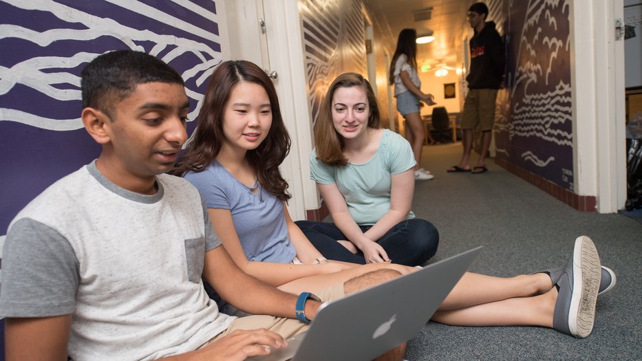 Students sit on floor looking at a computer in Lloyd House hallway