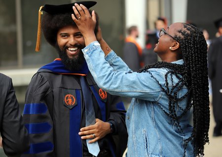 A woman with long dark braids and a denim jacket straightens the mortarboard of a smiling man who is graduating from Caltech