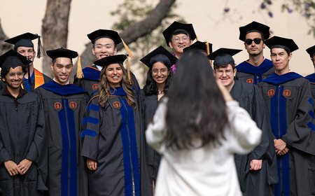A woman in a white jacket with long dark hair takes a photograph of 12 Caltech graduates
