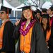 A group of graduates in caps and gowns processes into their commencement ceremony. One woman smiles at the camera and flashes the peace sign.