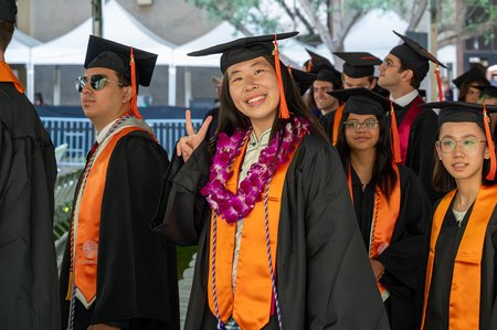 A group of graduates in caps and gowns processes into their commencement ceremony. One woman smiles at the camera and flashes the peace sign.