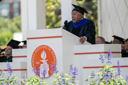 An older man wearing graduation robes with a blue stole speaks to an audience from a lecturn