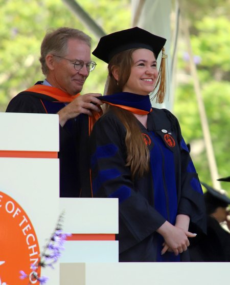 An older man wearing glasses places an orange stole around the neck of a woman who is graduating from Caltech