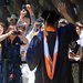 A man standing with his back to the camera wearing graduation robes waves to a group of people who are photographing him