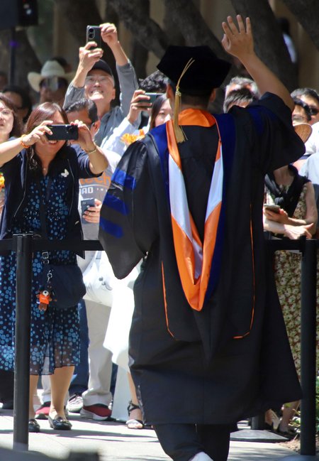A man standing with his back to the camera wearing graduation robes waves to a group of people who are photographing him