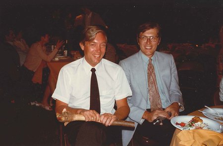 Two men sit at a table in a darkened room and smile at the camera. Other tables and partygoers can be seen sitting at tables in the background.