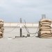 A sculptural installation consisting of a simple tripod rocket launcher with piles of sandbags on either side, and liquid fuel tanks on either side of the sandbags. The tanks and the launcher on the tripod are connected with many cables.