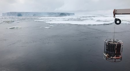 A large block of ice in the distance, ocean in the foreground. A cylindrical instrument hangs in frame at the lower right.