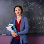 Image of Katerina Chatziioannou standing in front of chalk board holding books in grey blazer and burgandy blouse