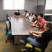 A person at a whiteboard talks to six people seated at a table and listening eagerly