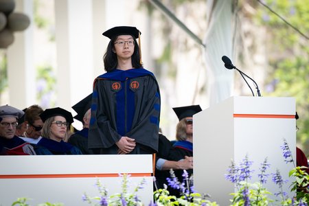 A young man with long dark hair and glasses dressed in his commencement robe stands as a commendation for him is read out.