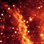 Astronomical image of the Double Helix Nebula in the Milky Way Galaxy. The nebula appears as two twisting ropes in orange surrounded by stars appearing as orange spots.