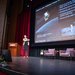 A speaker smiles and gestures onstage in front of a slide featuring the Lunar Trailblazer mission