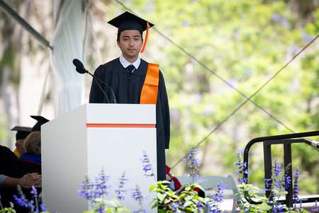A young man wearing a black robe and mortarboard with an orange tassle stands as a commendation for him is read out.