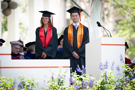 Two college graduates, both in graduation garb stand side by side. One is a young woman with a red sash and medium brown hair and the other is a young man with a lavender tie and short black hair.