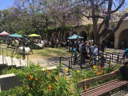 A crowd of families stands under trees and shade umbrellas on campus