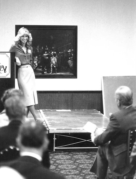 A black and white photo of a woman on an elevated stage at a lectern speaking before an audience.