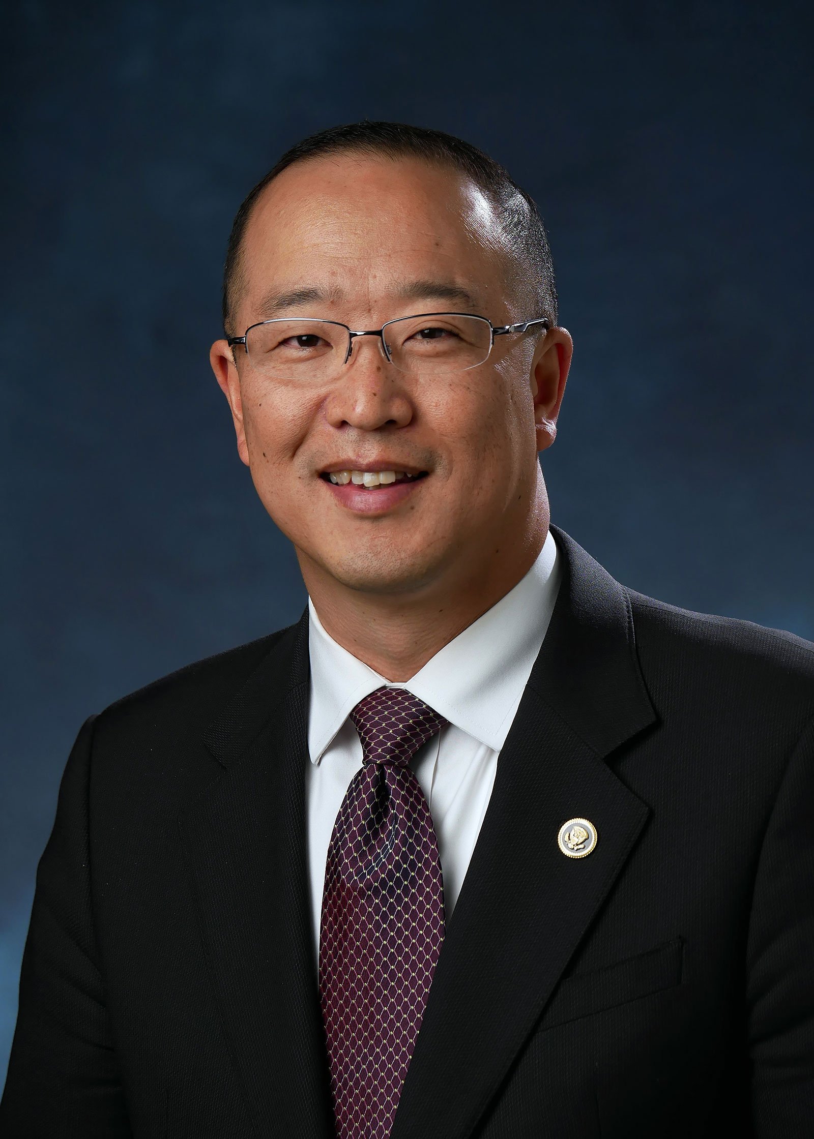 Portrait of David Kang wearing glasses and a dark suit and tie.