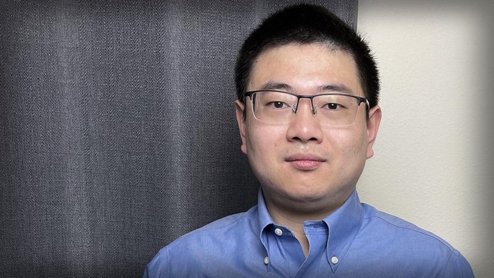 A portrait of Lei Li, who stands in his office wearing a button-down shirt and glasses.