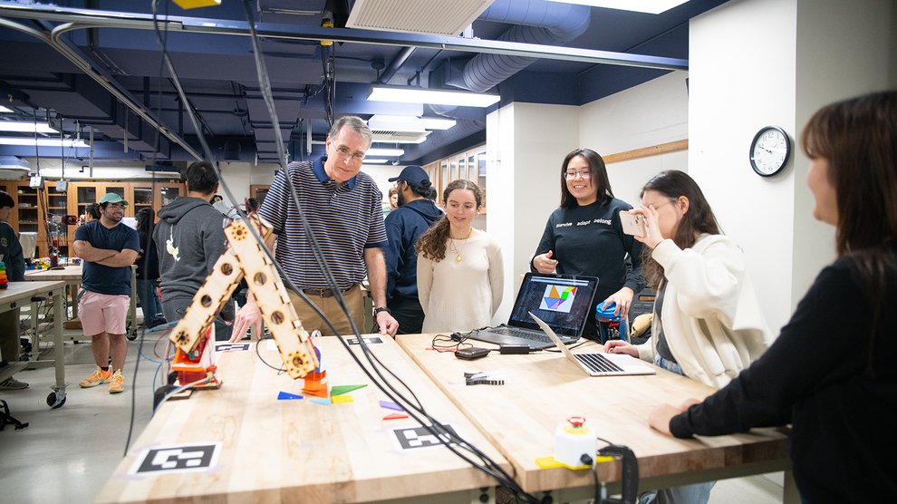 A robotic arm system is on a table, assembling a puzzle using tangrams. Several people are watching its actions. A man on the left is the course instructor, Gunter Niemeyer.