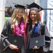 Two women in graduation robes stand next to one another holding their diplomas. The woman on the left has blond hair and a pink flowered lei; the woman on the right has shoulder-length medium brown hair and wears a blue sash.