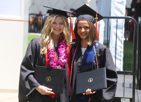 Two women in graduation robes stand next to one another holding their diplomas. The woman on the left has blond hair and a pink flowered lei; the woman on the right has shoulder-length medium brown hair and wears a blue sash.