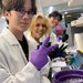 Four students are working in a scientific lab wearing rubber gloves, and using scientific instruments and a computer. All are smiling and facing the camera.