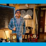 Rana Adhikari stands in his office wearing a plaid shirt and beanie, surrounded by clocks and maps and a chalkboard