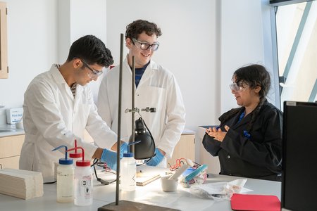 Two students in white lab coats on the left are interacting with a woman in a black lab coat on the right. The students are working on a lab using a simple solar cell.