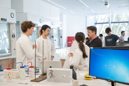 Three students in white lab coats are listening to an instructor in a black lab coat in a new white lab space.