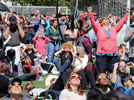 A densely gathered crowd, adorned with eclipse glasses, looks toward the sky in awe at the eclipse's peak at 11:13 a.m. in Pasadena. Amid the excitement, one person raises their arms in jubilation, while many others smile and cheer, visibly thrilled by the celestial spectacle.