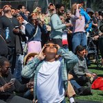 A large group of people wearing orange eclipse glasses gathered on Caltech's athletic fields. They are looking up toward the sky, observing an eclipse.