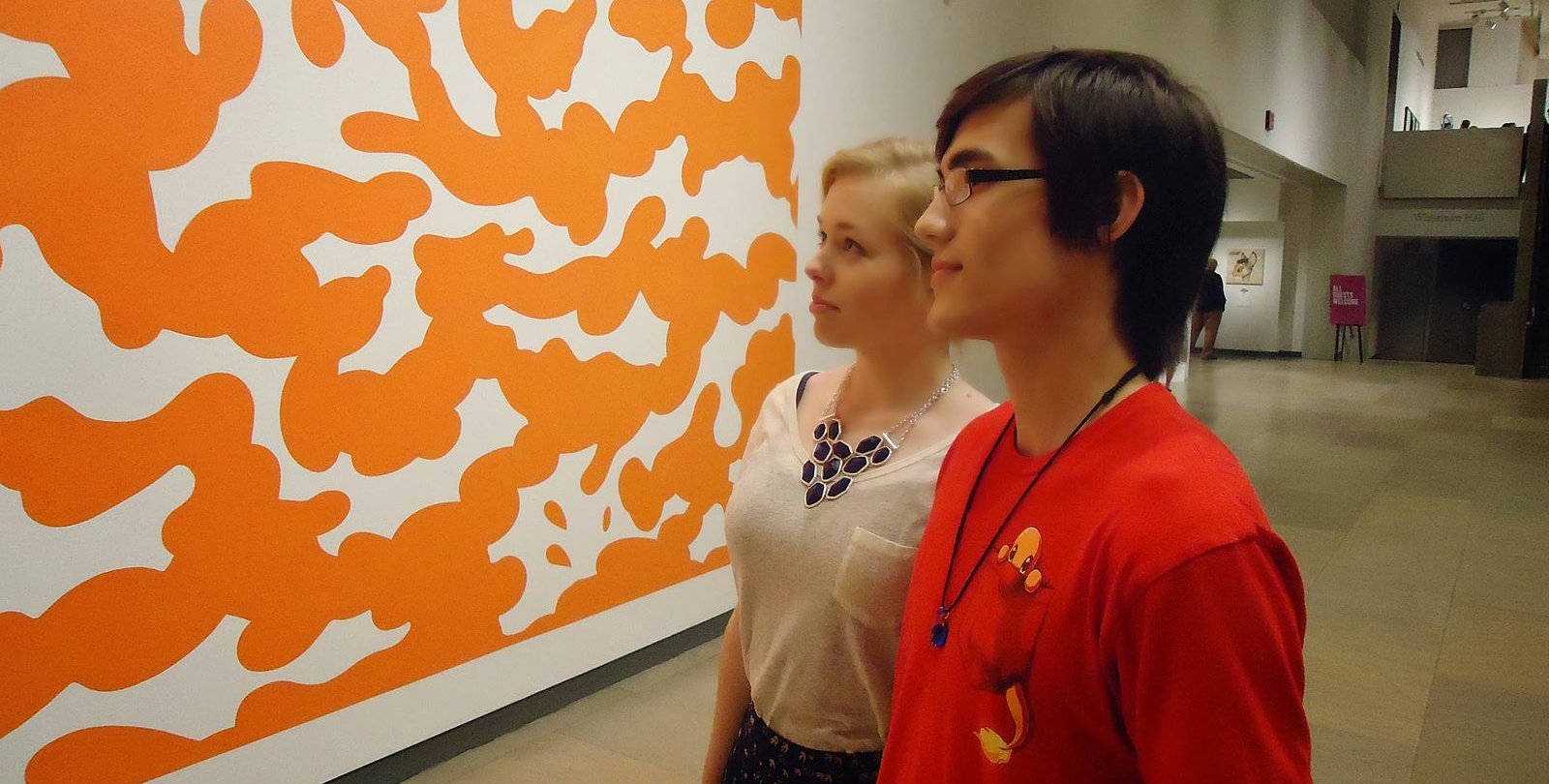 A couple looks at a piece of art in the Phoenix Art Museum. They are a young man and young woman who look at a modernist painting resembling orange clouds.