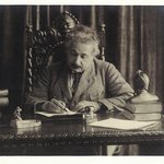 Einstein writes by hand at his desk. A bust of Dante sits on the corner of the desk near some books.